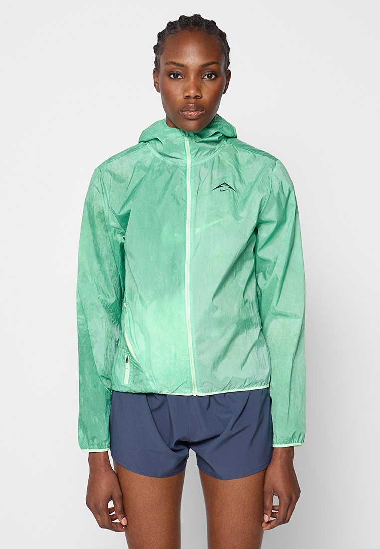Lekki, półprzezroczysty miętowy płaszcz z kapturem, z pełnym zamkiem z przodu i logo na lewej piersi. W połączeniu z ciemnoniebieskimi szortami.