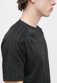 Μαύρο αθλητικό κοντομάνικο μπλουζάκι με τρεις κάθετες ρίγες στους ώμους, με στρογγυλή λαιμόκοψη και υφασμένα �σχέδια ραφών.