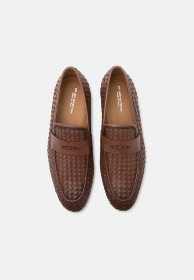 Καφέ υφαντά δερμάτινα loafers με διακοσμητικό λουράκι, με την ετικέτα Stuart Weitzman, κατασκευασμένα στην Ιταλία, εμφανίζονται από πάνω σε λευκό φόντο.