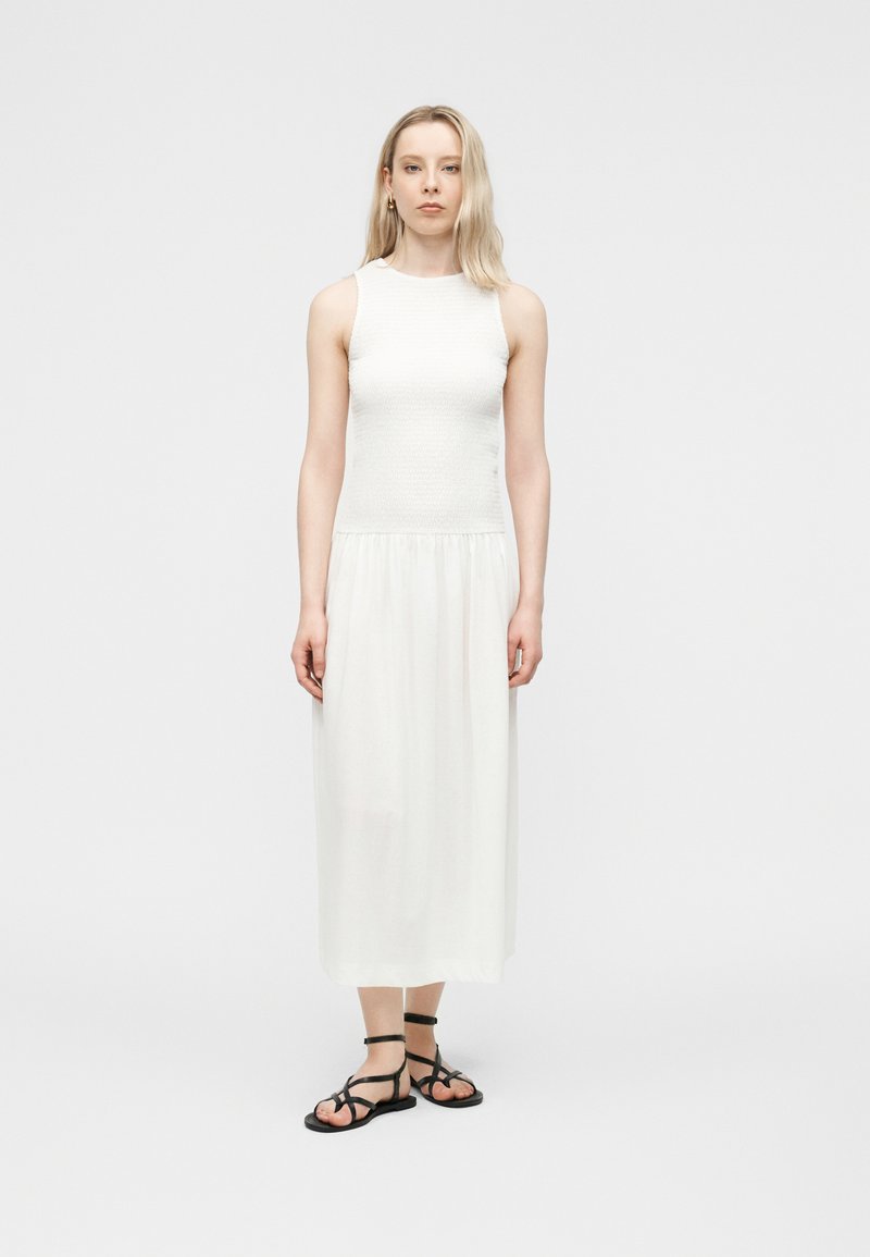 Γυναίκα με μακριά ξανθά μαλλιά, φορώντας λευκό φόρεμα χωρίς μανίκια και μαύρα πλατφόρμα σανδάλια με λουράκια, στέκεται μπροστά σε απλό λευκό φόντο.