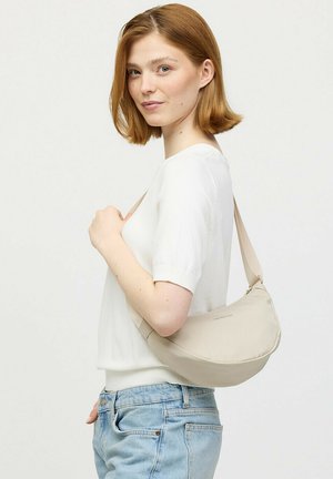 Jonge vrouw met rood haar die een wit shirt met korte mouwen en blauwe spijkerbroek draagt, houdend een beige schoudertas tegen een effen achtergrond.