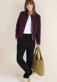 Tmavě fialová huňatá bunda přes bílý tričko, doplněná černými kalhotami na stažení a krémovými teniskami, držící olivově zelenou tašku.