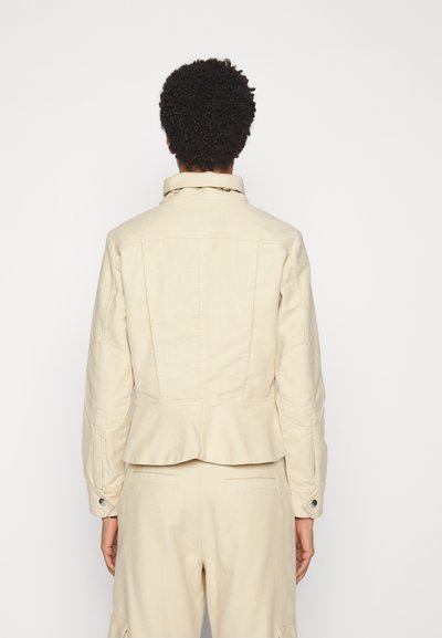 Beige cropped takki, joka on valmistettu puuvillasta ja jossa on kaulus, pitkät hihat sekä peplum-helma. Takana on näkyvää ompelua ja nappikoristeita.