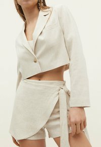 Beżowa lniana krótka marynarka zapinana na jeden guzik z naszywaną kieszenią, w połączeniu z dopasowanymi spódnico-spodniami wiązanymi w talii.