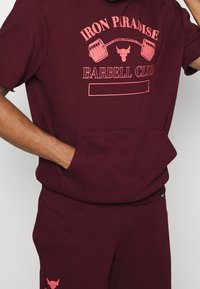 Sudadera burdeos de mangas cortas, con un gráfico en rosa de pesas y texto. Tiene un bolsillo tipo canguro y pantalones de chándal a juego con el logo.