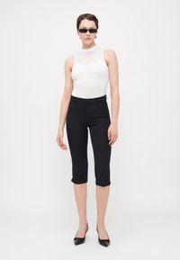 Juodos prigludusios capri kelnės derinamos su balta bodysuit, turinčia aukštą kaklą ir permatomą tinklinį intarpą. Modelis dėvi minimalistinius juodus akinius nuo saulės.