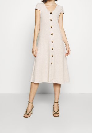 Femme portant une robe midi beige à manches courtes avec des boutons et un décolleté en V, ainsi que des sandales à talons hauts vert olive.