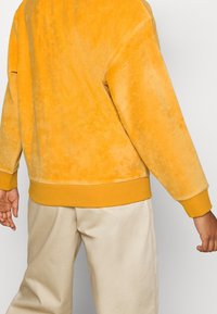 Felpa in velluto giallo con polsini e vita a coste, abbinata a pantaloni beige chiaro. Il tessuto ha una consistenza morbida e liscia.