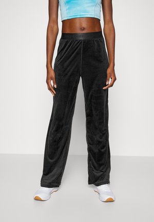 Osoba ubrana w czarne szerokie spodnie z sztruksu, jasnoniebieski krótki top na ramiączkach i białe trampki, stojąca na tle jednolitego tła.