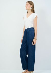 Camiseta blanca de cuello en v combinada con pantalones de mezclilla de pierna ancha en azul oscuro, con cintura ajustable y dos bolsillos frontales.