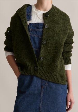 Personne portant un cardigan vert foncé boutonné sur une chemise blanche à manches longues côtelée et une salopette en denim bleu, avec une main dans la poche.