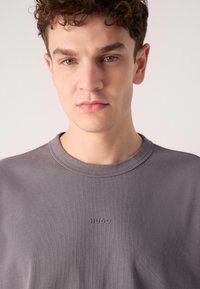 T-shirt gris à col rond en coton doux, avec un logo "HUGO" en relief subtil sur la poitrine. Texture lisse avec un design minimal.