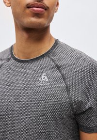 Άνδρας που φορά γκρι αθλητική κοντομάνικη μπλούζα με υφή, με λευκό λογότυπο Odlo στο στήθος, μπροστά σε απλό φόντο.