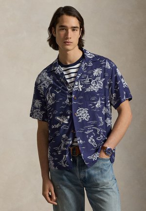 Νέος άνδρας που φοράει μπλε πουκάμισο Χαβάης με λευκά ναυτικά και τροπικά σχέδια, ριγέ εσωτερική μπλούζα, μπλε τζιν και ρολόι χειρός.