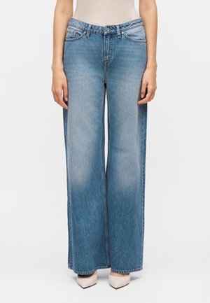 Kobieta ubrana w jasnoniebieskie szerokie dżinsy i kremowe buty na obcasie z ostrym noskiem, stojąca na jednolitym tle.