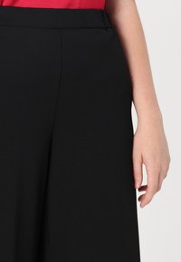 Mustad laiad püksid siledast materjalist, millel on lame eesmine vöökoht ja lõdvestunud siluett, osaliselt näidatud käe abil.