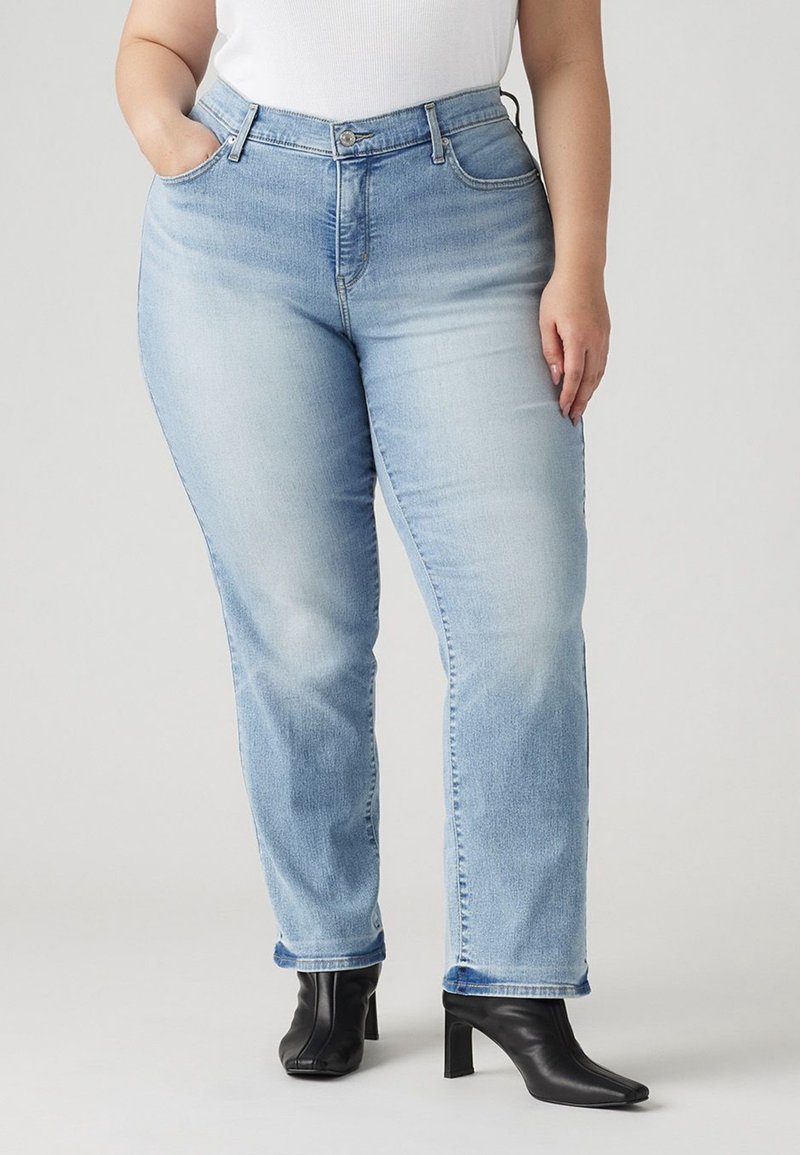 Femme portant un jean droit bleu clair, un haut blanc et des bottines à talons noires, avec une main dans la poche, sur un fond uni.