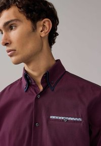 Camisa burdeos de botones con un tejido texturizado, cuello en contraste azul marino y un detalle de bolsillo con patrón.
