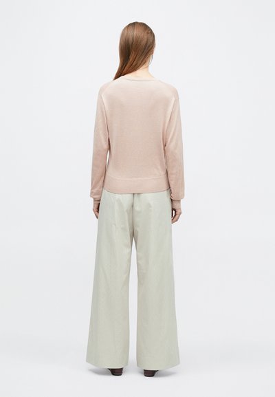 Frau mit langen Haaren, die einen hellrosa Pullover und weite beige Hosen trägt, steht mit dem Rücken zur Kamera vor einem einfarbigen Hintergrund.