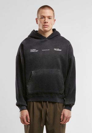 Black cotton hoodie with a front pocket, loose fit, and oversized sleeves. Features white printed logos on the chest and sleeve.