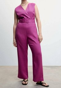 Salopette en lin fuchsia avec un devant enveloppant, des jambes larges et une taille ceinturée. Le mannequin porte des tongs noires et un collier en chaîne dorée.