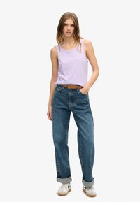 Lichtpaarse mouwloze tanktop, high-waisted blauwe jeans met opgerolde boorden, bruine riem, witte en grijze sneakers, staand tegen een effen achtergrond.