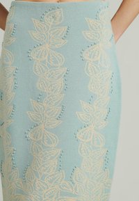 Helle blaue Bleistiftrock mit beigem Blätter-Stickerei und kleinen Perlenakzenten. Der Stoff hat eine glatte Textur und eine taillierte Silhouette.