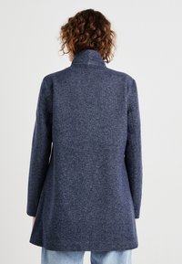 Marineblauer, melierter Mantel mit hohem Kragen, langen Ärmeln und ausgestelltem Schnitt, gefertigt aus einem weichen, strukturierten Stoff.