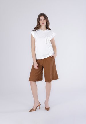 Donna in piedi con una mano nei pantaloncini marroni fino al ginocchio e top bianco senza maniche con tasca decorativa, indossa scarpe con tacchi alti marroni.