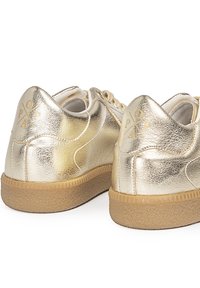 Zapatillas metálicas doradas con un acabado texturizado, suela de goma marrón contrastante y detalle del logotipo en el talón. Presenta cordones y una forma redondeada.