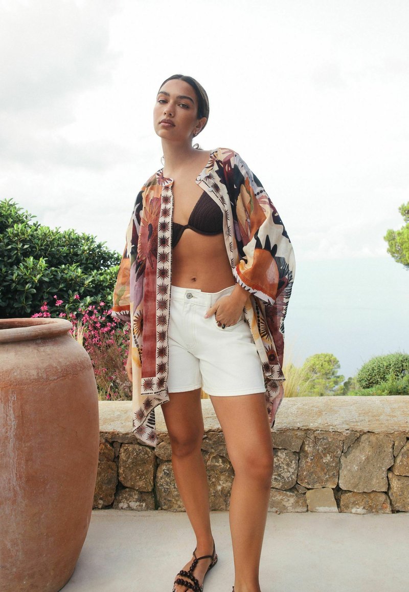 Letvægts blomstret kimono med livlige mønstre, kombineret med en brun bikini, hvide shorts og sorte sandaler, sat mod en naturlig baggrund.