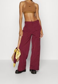 Cargo byxor i djup bourgogne, med lös passform, två framfickor och knapplåsning. Kombineras med en brun Nike crop top och en väska med gula accenter.