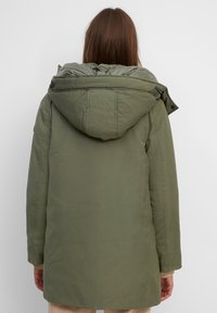Giacca parka isolata verde oliva con cappuccio. Presenta cuciture visibili e due tasche laterali, realizzata in materiale durevole e resistente all'acqua.