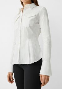 Woman wearing a fitted white ribbed button-down shirt with front pockets and flared cuffs paired with black pants.