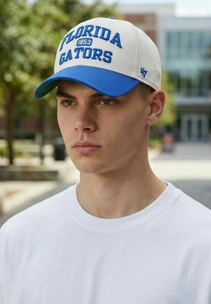 Junger Mann im Freien, trägt eine weiße und blaue Baseballkappe der Florida Gators und ein schlichtes weißes T-Shirt, mit verschwommenem Hintergrund aus Bäumen und einem Gebäude.