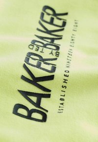 Jasnożółty materiał z czarnym wytłoczonym napisem "BAKER BAKER" z napisem "ZAŁOŻONE JEDENAŚCIE OSIEMDZIESIĄT OSIEM" poniżej. Miękka tekstura.