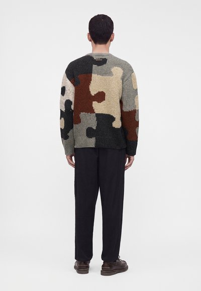 Мъж, облечен с пуловер с модели на многоцветни пъзелови парчета, черен панталон и кафяви обувки, стоящ с гръб към камерата на обикновен фон.