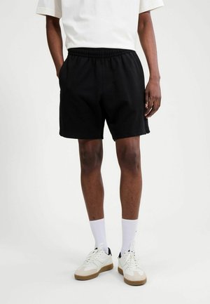 Person trägt schwarze Shorts mit elastischem Bund, weiße Sportsocken, weiße Turnschuhe mit Gummisohlen und ein weißes kurzärmliges Shirt und steht vor einem einfarbigen Hintergrund.