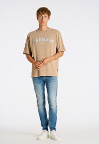 Beige puuvillane t-särk, millel on "LINDBERGH 1962" trükk, koos heledate siniste teksade ja valgete spordijalatsitega, seismas ühetoonilise tausta ees.