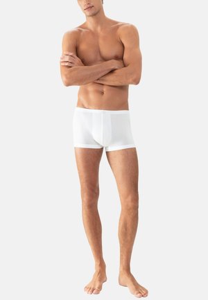 Mann, der barfuß mit verschränkten Armen steht, bekleidet mit weißen eng anliegenden Boxer-Shorts vor einem schlichten hellen Hintergrund.
