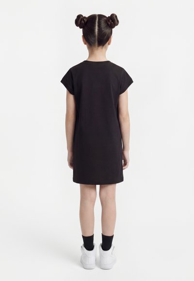 Παιδί που κοιτάζει προς τα πίσω, φοράει μαύρο φόρεμα με κοντά μανίκια, μαύρες κάλτσες, λευκά αθλητικά παπούτσια, με τα μαλλιά του κομμένα σε δύο κότσους.