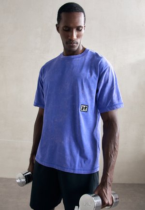 T-shirt en coton violet à manches courtes et col rond, avec un petit patch logo. Le modèle tient des haltères en acier inoxydable.