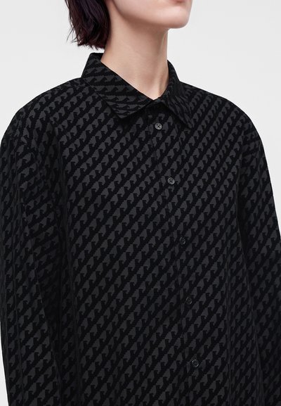 Personne portant une chemise noire à boutons avec un motif pied-de-poule diagonal texturé et un large col.