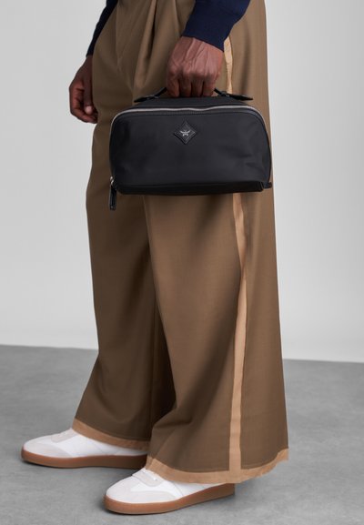 Czarna torba na kosmetyki z tkaniny, z dwoma uchwytami, srebrnym zamkiem błyskawicznym i naszywką w kształcie diamentu, trzymana przez osobę ubraną w beżowe spodnie w szerokim kroju i białe trampki.