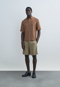 Homme debout portant une chemise à manches courtes marron à motifs avec boutons, un short beige à motifs et des chaussures habillées noires, sur un fond uni.