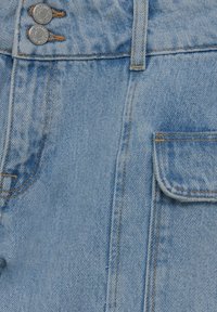 Des jeans en denim bleu clair présentent deux boutons métalliques, des coutures oranges et une poche latérale avec un rabat arrondi.