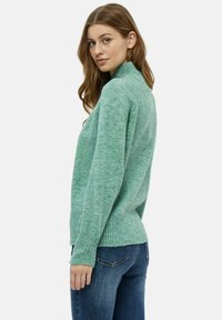 Maglione lavorato a maglia verde con collo alto, orlo a costine e maniche lunghe, abbinato a jeans in denim blu; texture morbida, vestibilità casual.