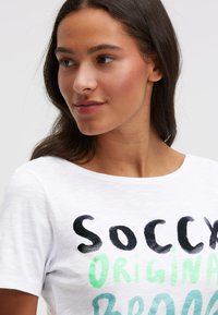 Weißes Baumwoll-T-Shirt mit dem Schriftzug "SOCCY ORIGINAL" in Schwarz und grünem Ombre. Rundhalsausschnitt, kurze Ärmel und weiche Textur.