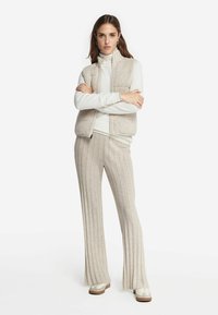 Beżowy sweter z dzianiny z dwoma przednimi kieszeniami, noszony na białą bluzkę z długim rękawem. Szerokie beżowe spodnie prążkowane dopełniają stylizację, w połączeniu z jasnymi sneakersami.