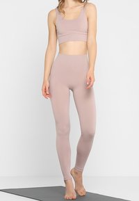 Kvinna som står barfota på en yogamatta iförd en ljus mauve sömlös sportbh och högmidjade leggings mot en enfärgad bakgrund.